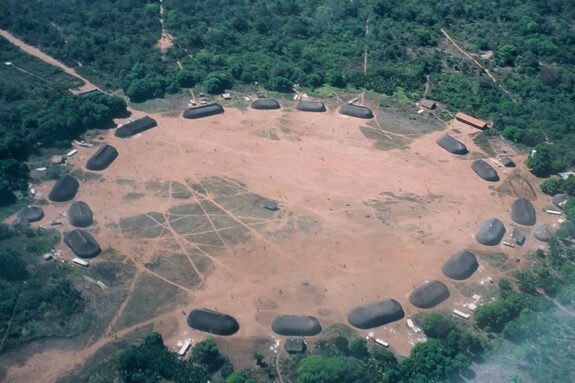 An Amazonian village with about 20 huts that form a large a ring around an empty, brown, circular area. Lots of trees are around the village.