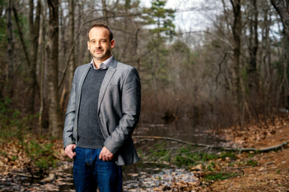 Greg Fournier stands in the woods, and there is a stream in background. There are fallen leaves and branches, and it is a cloudy day in early Spring.