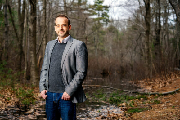 Greg Fournier stands in the woods, and there is a stream in background. There are fallen leaves and branches, and it is a cloudy day in early Spring.