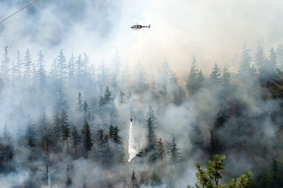 A helicopter drops water on a vast, smokey forest fire
