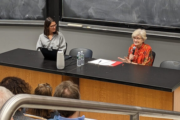 Kate Zernike and Nancy Hopkins sit at a table addressing an audience in a classroom. A blackboard appears behind them.