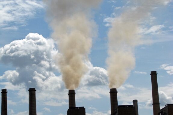 Photo of a row of smokestacks. Two are belching smoke into a cloudy blue sky.