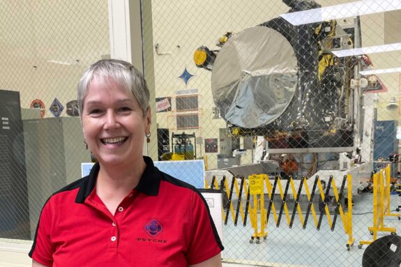 Lindy Elkins-Tanton poses in a red, collared Psyche shirt. A large silver disc on the spacecraft can be seen through the window behind her.