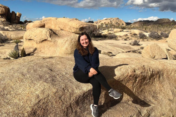 Becca Mastrola sits on a rock formation and smiles at the camera.