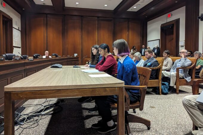 Erin Cusson, Prajna Nair, and Ella Sheffield sit at a table giving testimony before a Massachusetts committee meeting.
