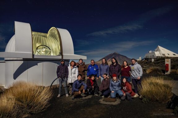 A group of 13 people pose at night next to a small domed telescope observatory. Mount Teide is visible behind them.