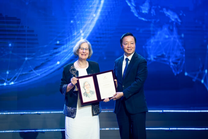 Susan Solomon and Tran Hong Ha stand on a stage in front of a blue background. They are both holding a folio open for the audience to see, and Solomon is holding a gold trophy.