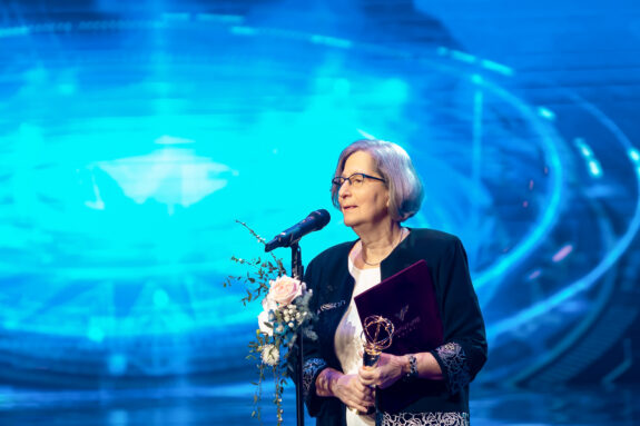 Susan Solomon stands on a stage in front of a blue background with a microphone. She is giving an acceptance speech and is holding a gold trophy in one hand.