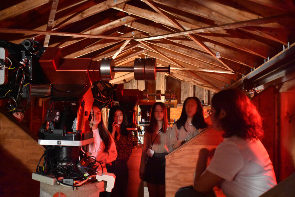 A group of five students in a wooden shed with red lighting. A telescope is in the foreground.
