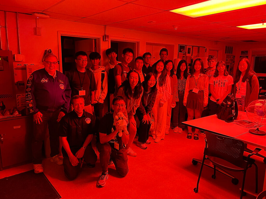 A group photo of 19 people inside the MIT Wallace Observatory in red light.