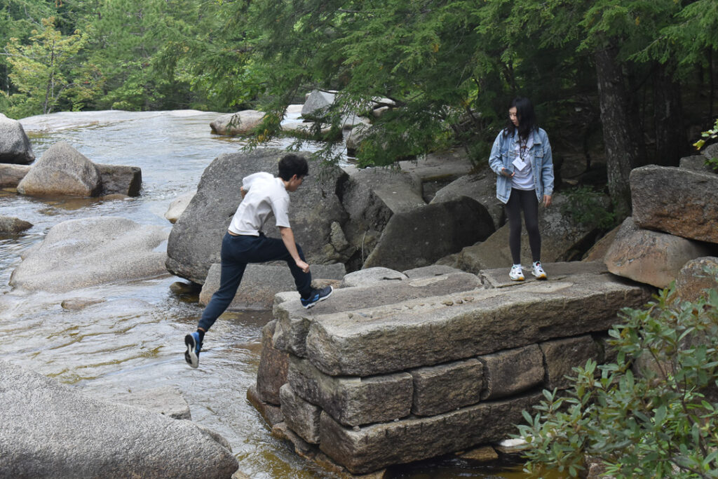 A person jumps from a rock in the middle of a river onto a stone outcrop, where another person is waiting for them.