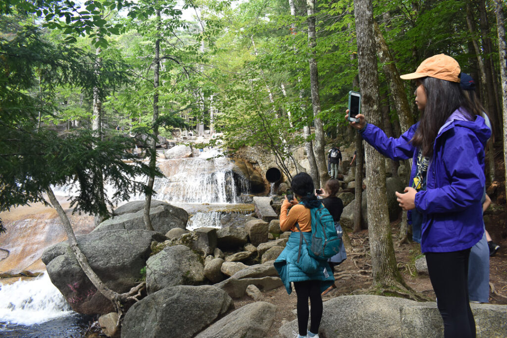 Three people stand on rocks on the edge of a river running through a forest, taking photos of a waterfall.