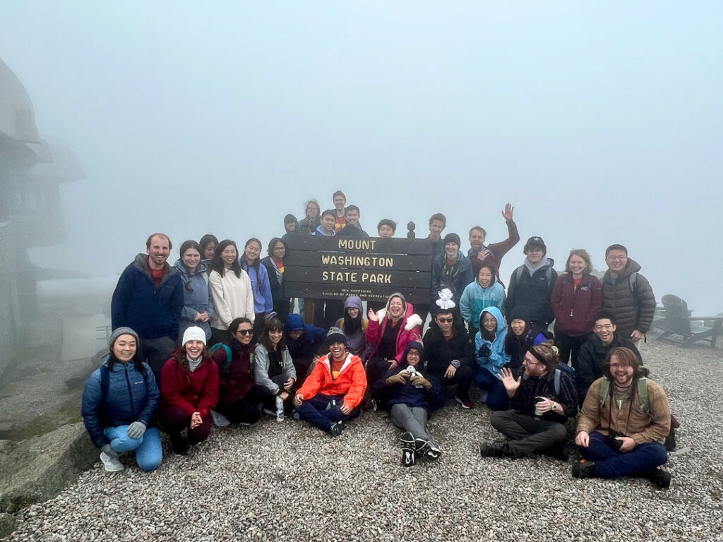 34 people pose for a photo at the top of a foggy mountain around a wooden sign that reads "Mount Washington State Park"