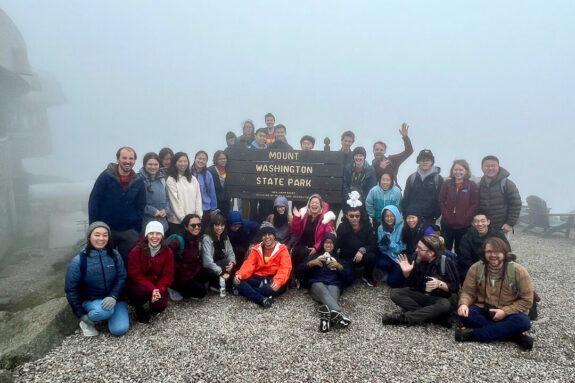 34 people pose for a photo at the top of a foggy mountain around a wooden sign that reads 
