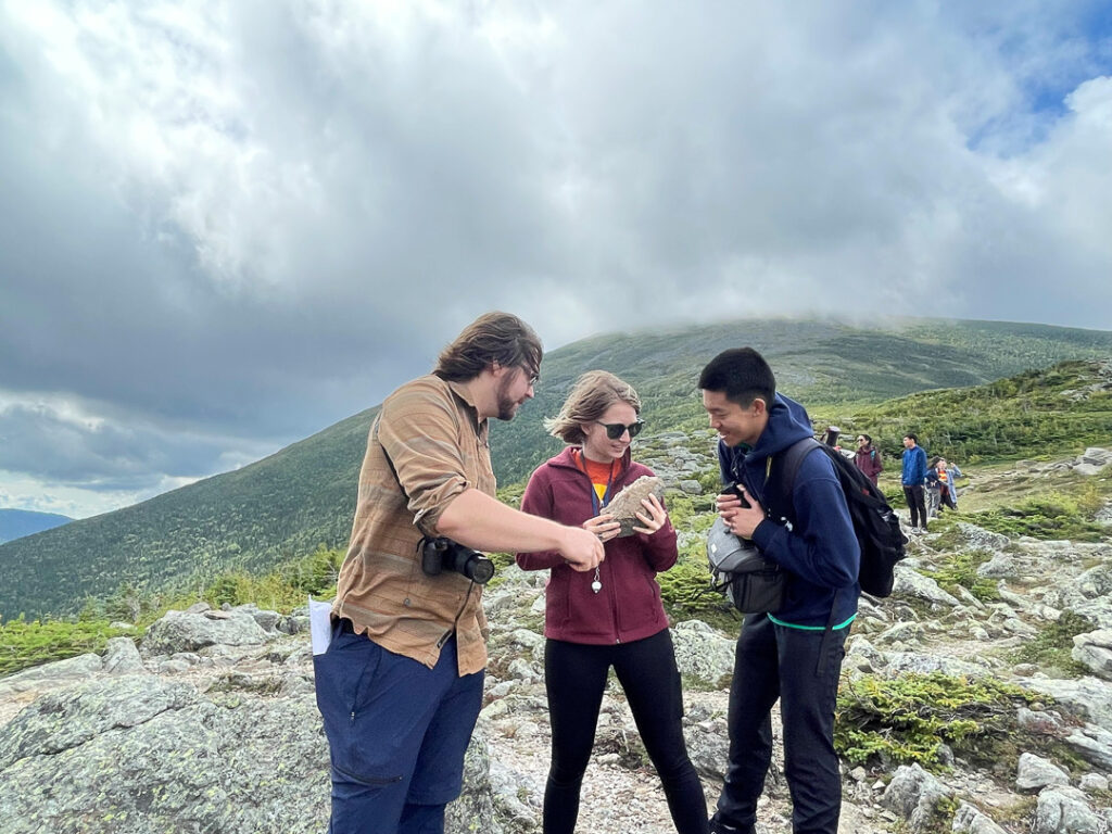 Three people stand together in a group looking at a large rock in one person's hand. They are on top of a mountain on a cloudy day.