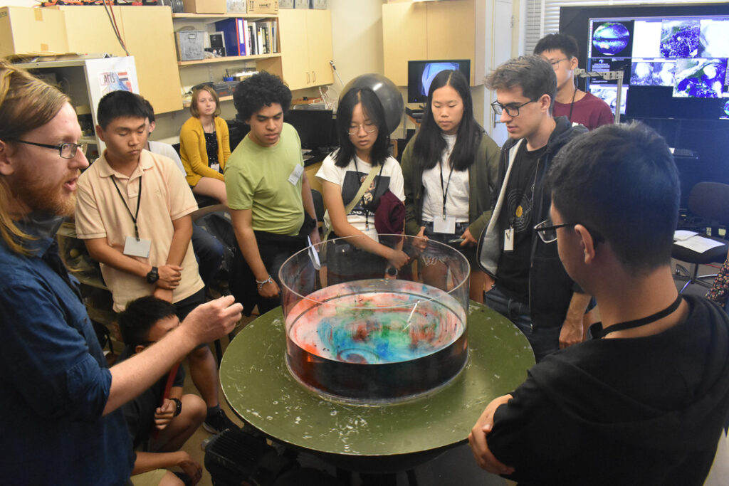 A group of students gathered around a table with a rotating tank filled with a clear fluid that has blue, red and green dyes mixed in it. One person in the group is talking.