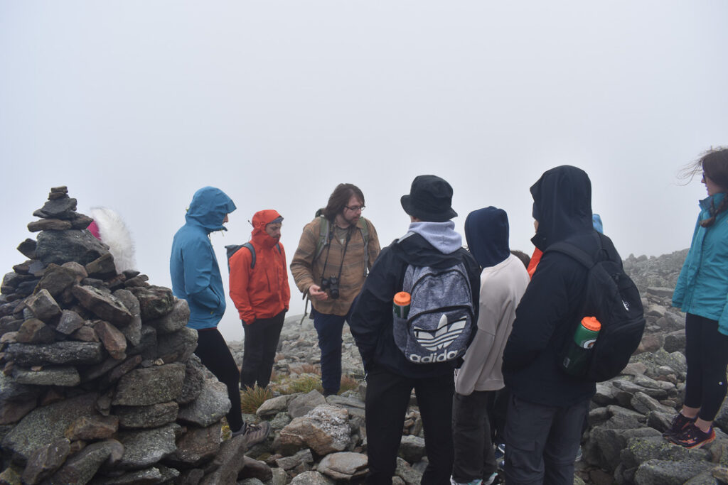 Seven people in rain coats stand on the edge of a rocky mountain in intense fog. Next to them is a stone cairn.