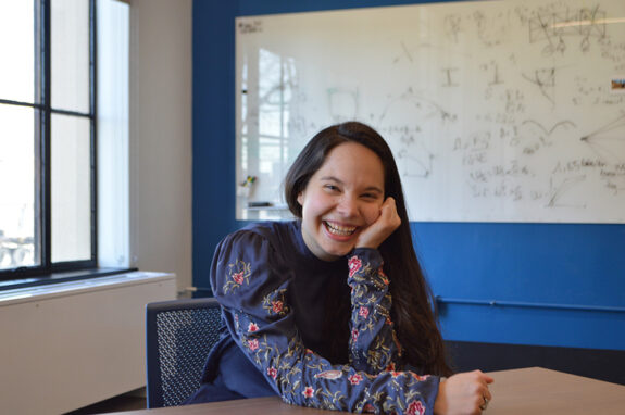 Ananda Santos Figueiredo sits at a table in front of a white board with math equations on it, smiling at the camera.