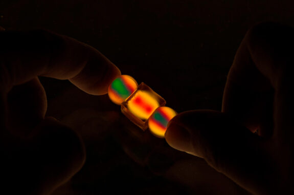 In darkness, thumbs hold two small spherical and one small cube objects that light up in vivid yellow, red, and green.