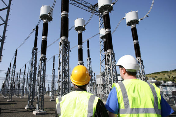 Two men in hardhats and safety vests, seen from behind, inspect a forest of electrical pylons and wires on a cloudless day.