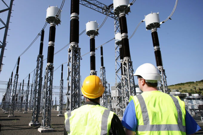 Two men in hardhats and safety vests, seen from behind, inspect a forest of electrical pylons and wires on a cloudless day.