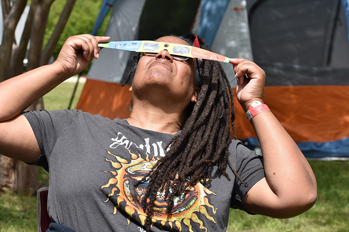 A photo of MIT alum Dara Norman with both hands holding a strip of material over her eyes while gazing up into the sky, with grass and a tent behind her.