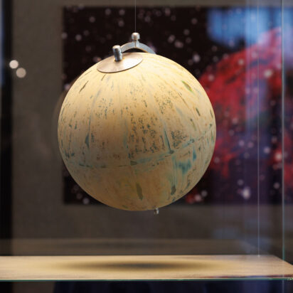 A globe that has been sandblasted to remove all distinguishing features hangs suspended in a square glass enclosure.