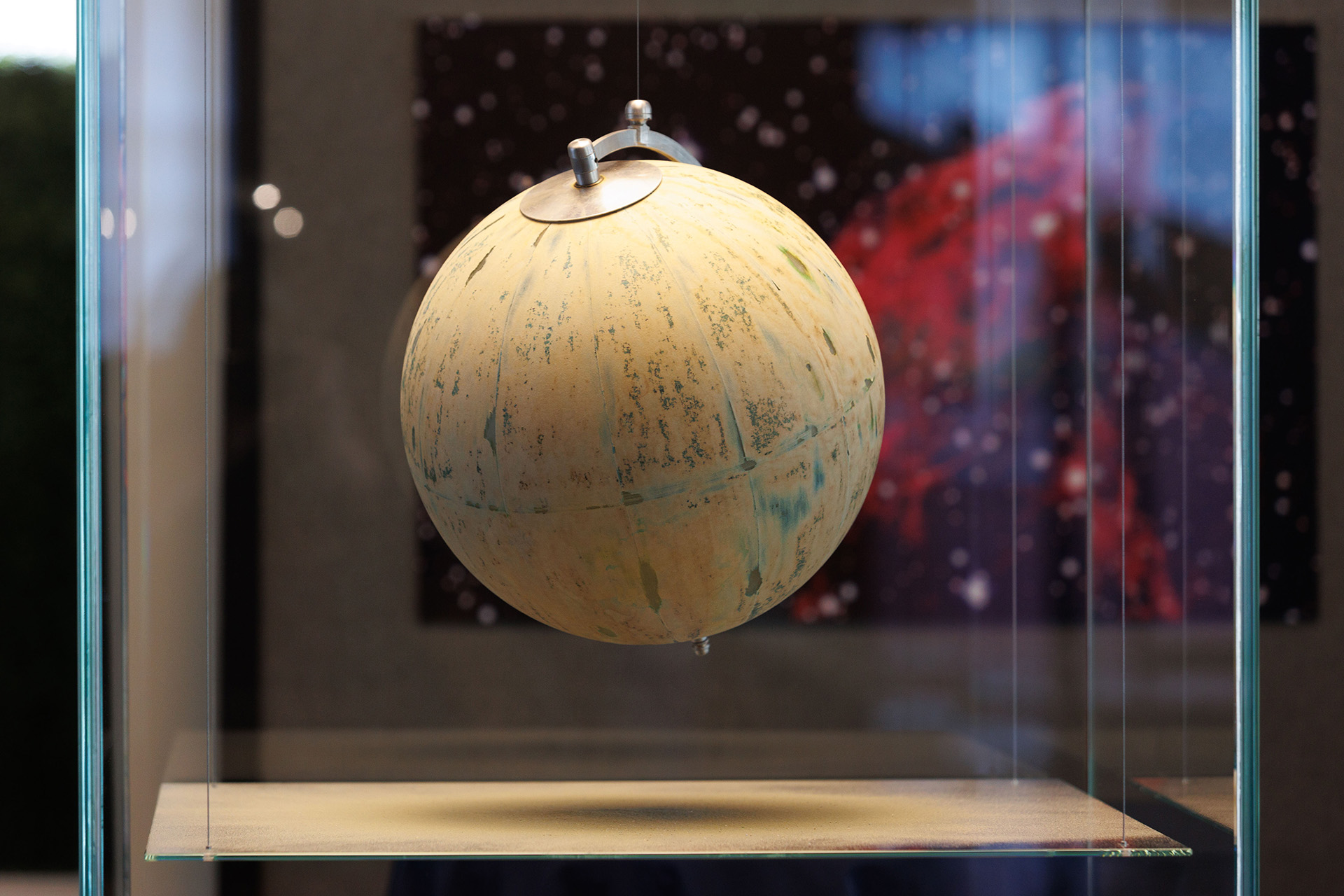 A globe that has been sandblasted to remove all distinguishing features hangs suspended in a square glass enclosure.