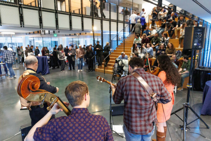 Four-person bluegrass band, seen from behind, performs in the Building 55 atrium with an audience of students, alumni, and faculty seated on tiered seating.