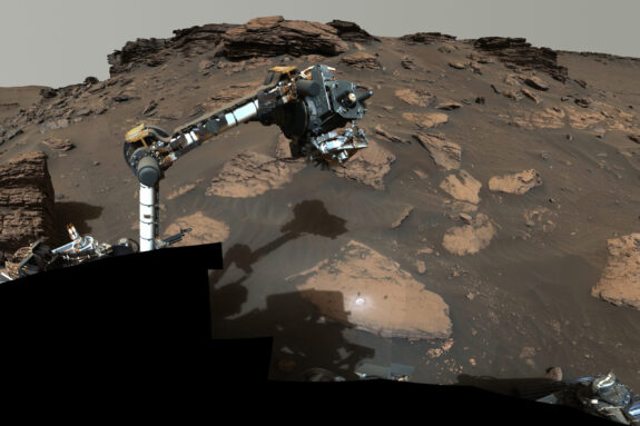 Photo shows the rover’s long arm and the rocky brown Martian soil.