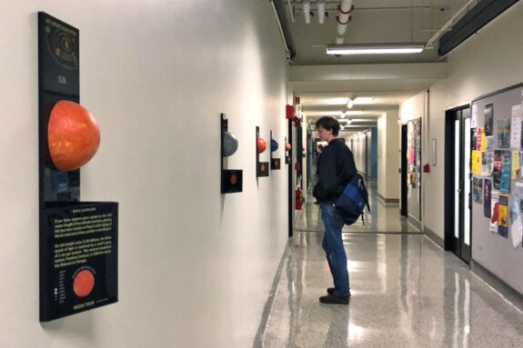 A student stands in front of palques depicting the Sun and inner rocky planets in 3D, positioned to scale in the long Infinite Corridor.