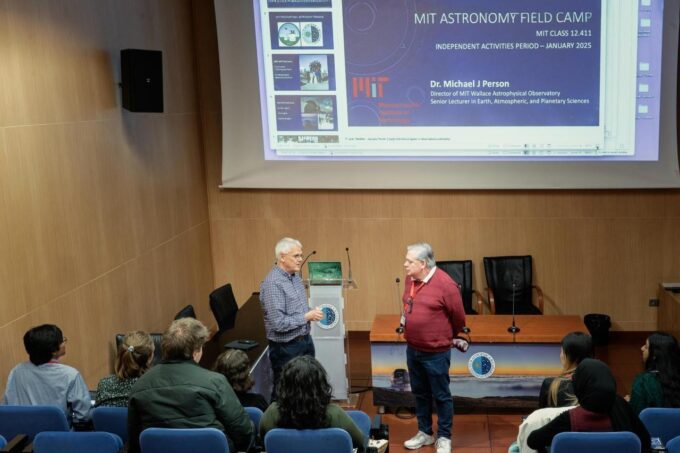 Michael Person stands at the front of a small lecture hall with a presentation behind him about the 2025 MIT Astronomy Field Camp.