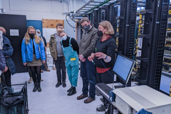 Seven masked individuals stand watch someone talking. They are standing near a computer in a space filled with wired fiber optic data hub.