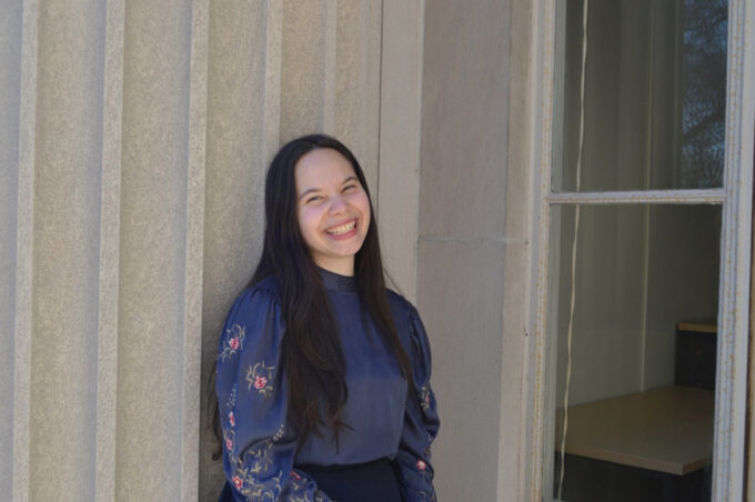 Ananda Santos Figueiredo leans against concrete building column.