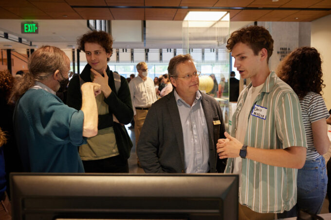 Four people standing in front of a computer monitor are talking with each other.