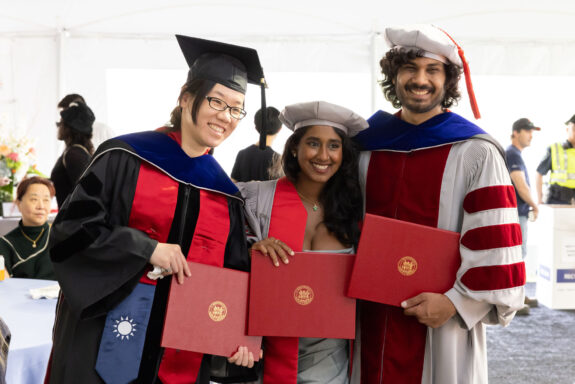 Three MIT PhD graduates in caps and robes pose with their diplomas and new doctoral hoods.