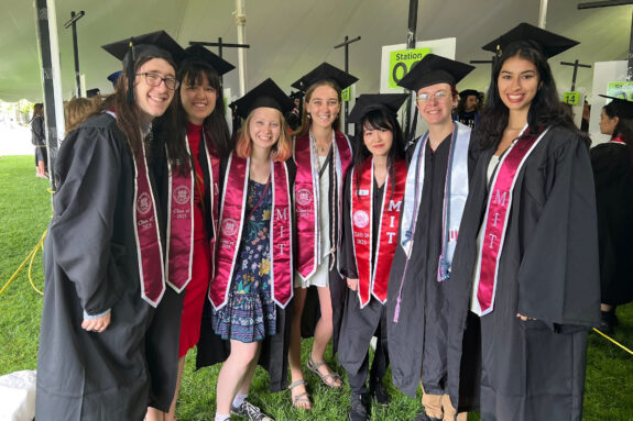 Seven from the EAPS Class of 2025 pose under a tent wearing their graduation caps and gowns.
