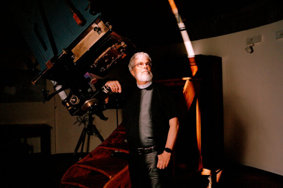 A photo of a man in religious attire leaning against a telescope.