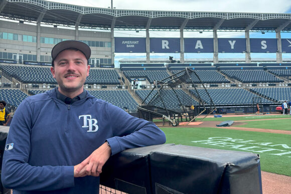 Michael McClellan poses in an empty baseball stadium. He is wearing a ballcap and has TB on his sweatshirt. The home plate area can be seen behind him and the stands are behind that. The word RAYS is spelled out behind the seats.