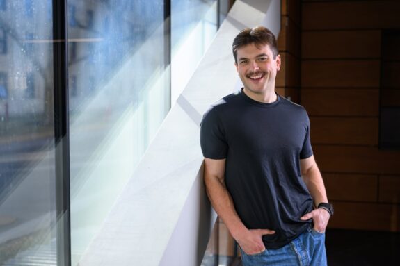 Perrin Davidson stands, posing, against an indoor angled pillar.
