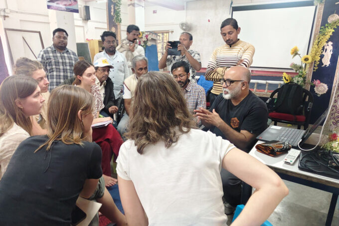 14 people, some holding notebooks, sit or stand in a collective huddle indoors as Sai Ravela explains how to play a game he created.