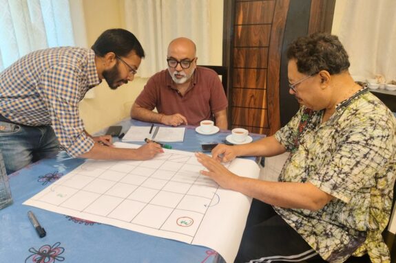 Sai Ravela, Anamitra Saha, and Jayanta Basu sit at a table with markers, drawing symbols onto a large piece of paper lined with a grid.